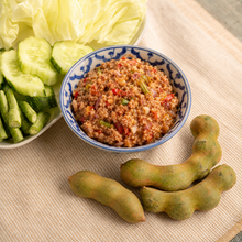 A bowl of prepared food with a blue floral pattern, served with sliced cucumbers and lettuce on a woven beige tablecloth, with three young tamarind pods in the foreground.