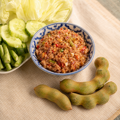 A bowl of prepared food with a blue floral pattern, served with sliced cucumbers and lettuce on a woven beige tablecloth, with three young tamarind pods in the foreground.