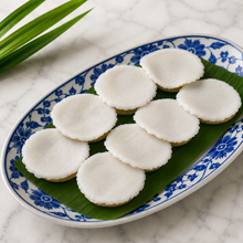 Eight round white cake slices, thai khanom tuay arranged on a green banana leaf inside an oval white ceramic plate with a blue floral pattern, set on a white marble surface.