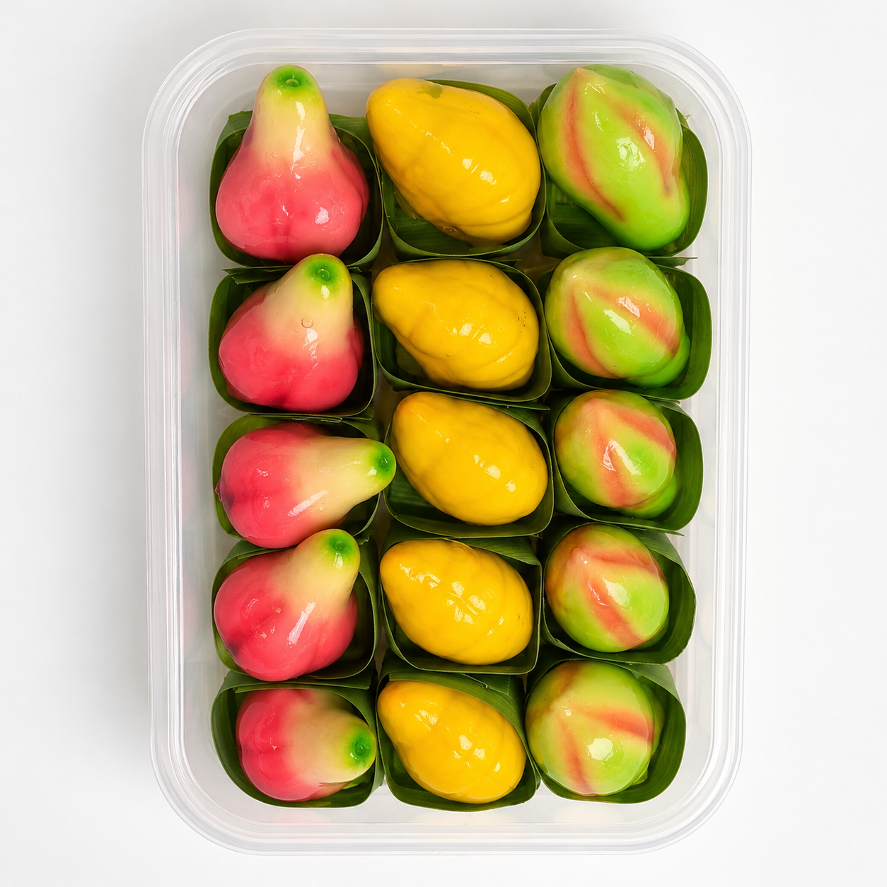 A clear plastic container holding 12 Thai fruit paste candies shaped like mangoes and lychees, arranged in two rows of six. The candies feature vibrant yellow, pink, and green colors, with some pieces showing a marbled pattern.